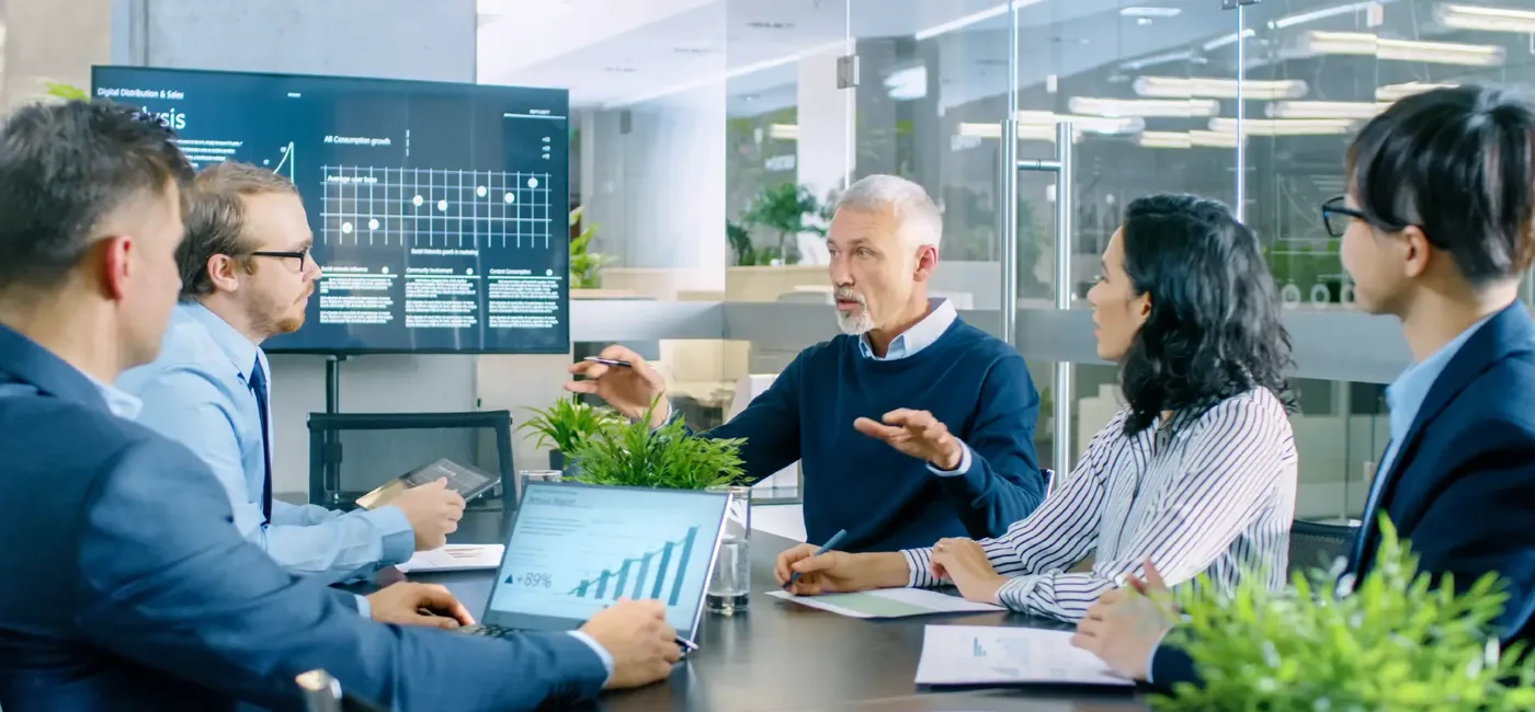 Colleagues in a glass-walled meeting room discussing charts on a screen