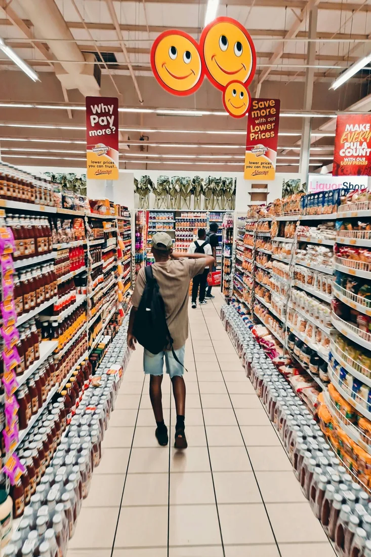 A shopper with a backpack walking down a busy supermarket aisle