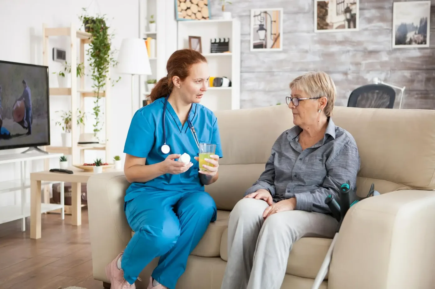 A nurse in blue scrubs talking with an older woman on a sofa